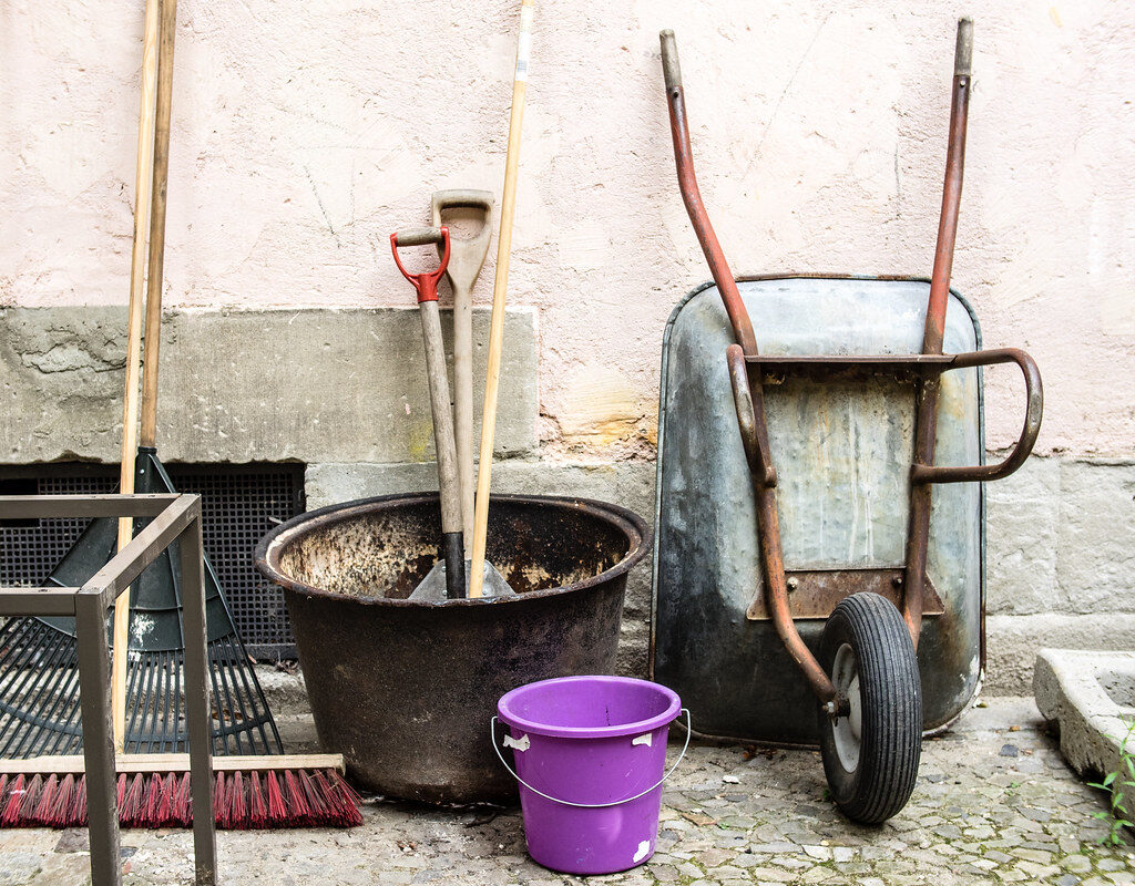 Wheelbarrow, rakes, brooms and bucket lined up against a wall ready for garden work
