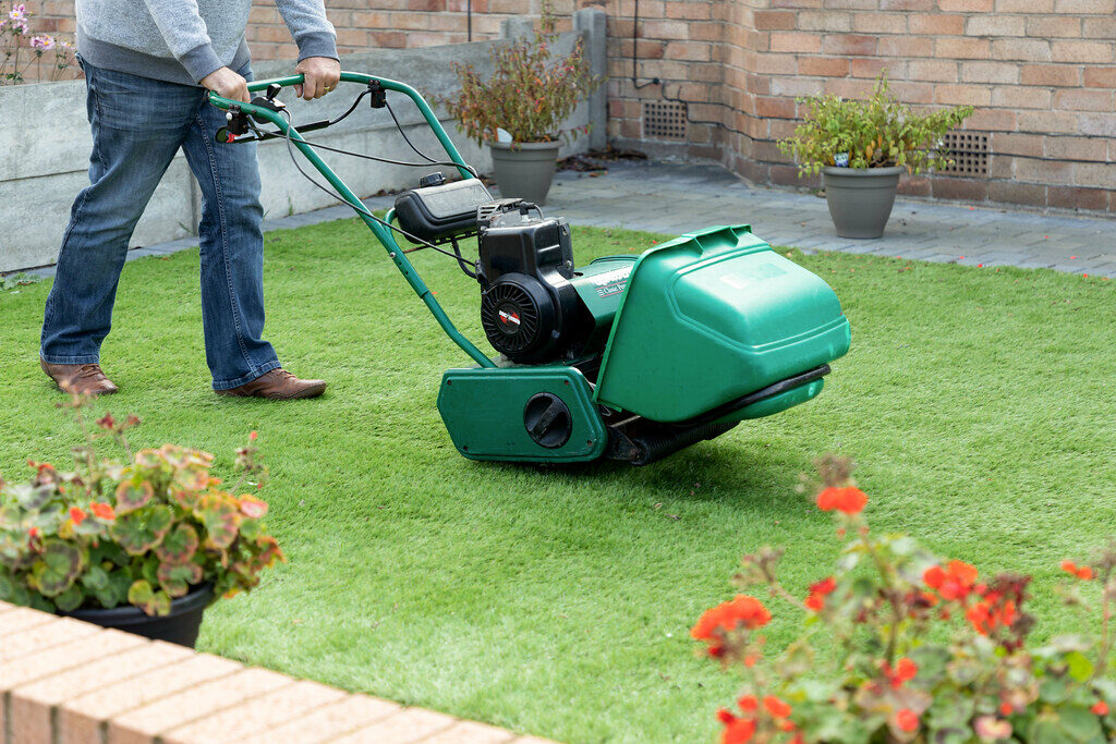 Man pushing a green petrol lawn mower across a freshly striped home lawn beside a brick house