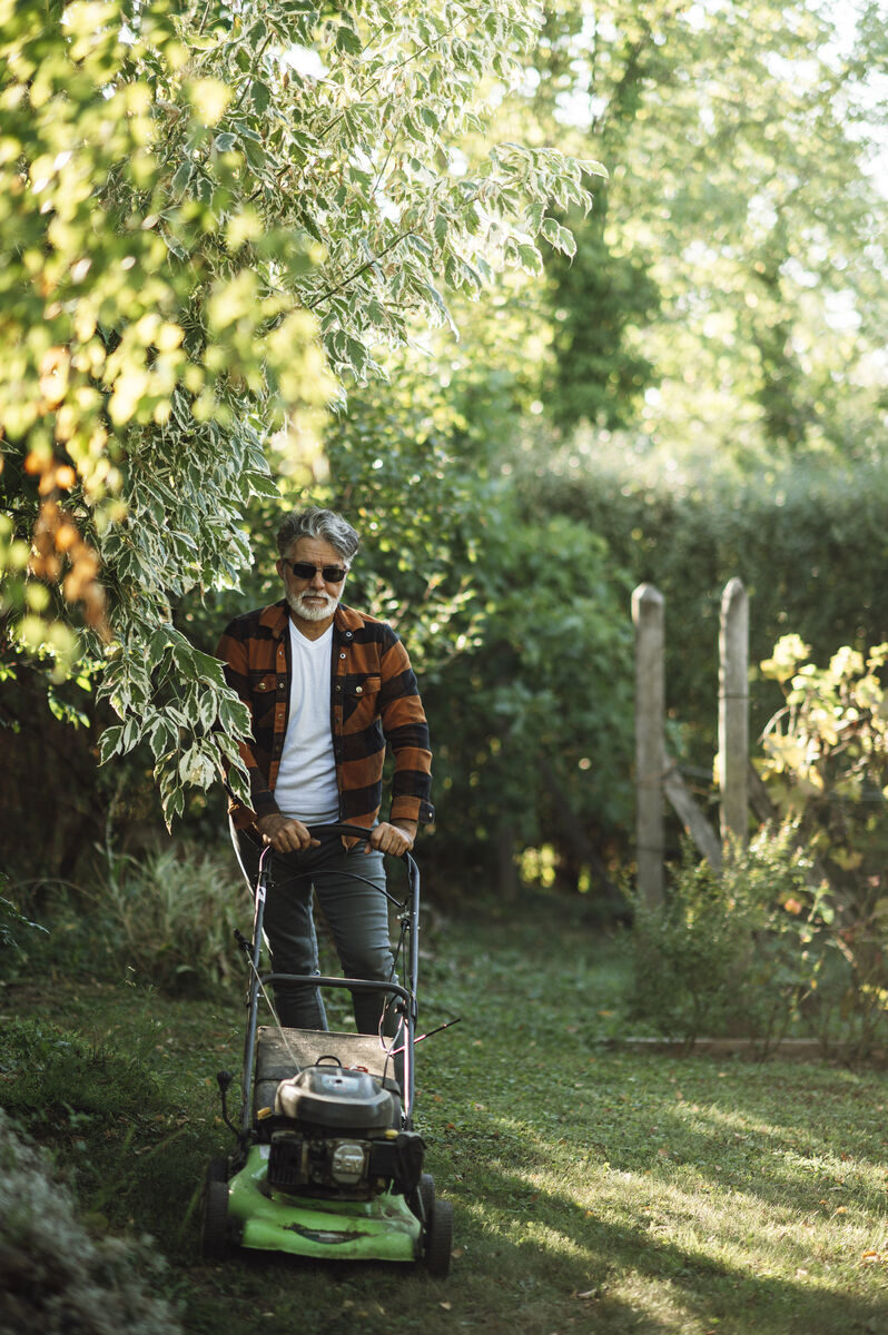 Man pushing a mower through a leafy shaded home garden on a sunny day